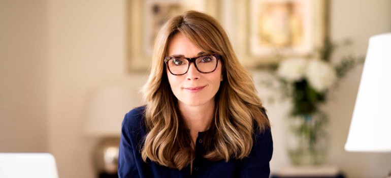 Eine Dame mittlere Alters mit Brille sitzt am Schreibtisch und lächelt