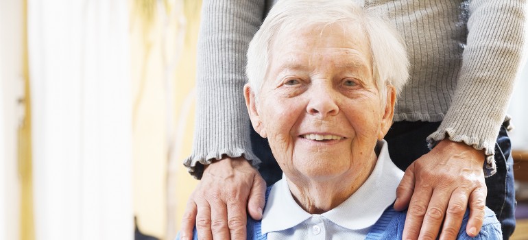 Eine ältere Dame sitzt in einem Stuhl, jemand hat von hinten die Hände auf ihre Schultern gelegt