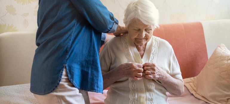 Eine ältere Dame knöpft sich in Anwesenheit einer Pflegeperson die Knöpfe ihrer Bluse zu